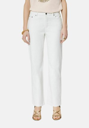 Kobieta ubrana w białe dżinsy o prostych nogawkach, jasnobeżową bluzkę, złote sandały na obcasie z paskami i złotą biżuterię na obu rękach na białym tle.