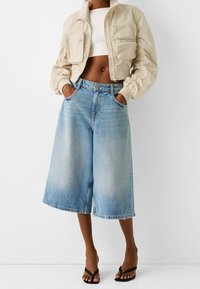 Beige cropped jacket with zippered pockets, paired with light blue wide-leg jeans. Black flip-flops complete the outfit.