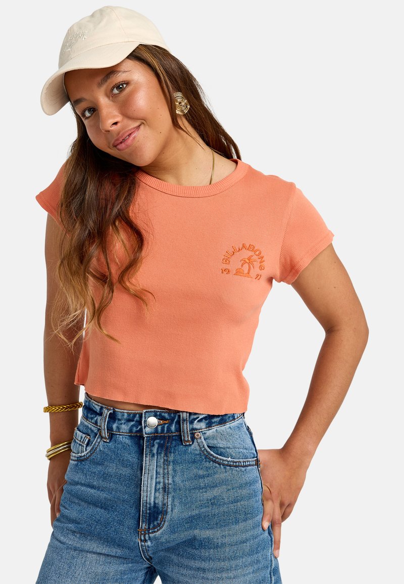 Cropped top a maniche corte e costine color pesca con logo ricamato. Abbinato a jeans in denim blu a vita alta. Cappellino beige e accessori in oro.
