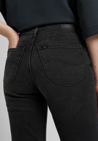 Black denim jeans featuring a mid-rise waistband, subtle stitching details, and a logo tag on the back pocket. Smooth texture and tailored fit.