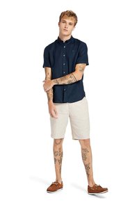 Camicia button-up a maniche corte di colore blu navy con colletto, indossata con pantaloni corti beige chiaro e mocassini marroni. Presenta tatuaggi visibili sulle braccia.