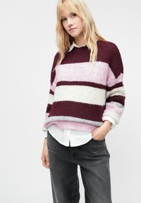 Pulôver de malha com riscas em bordeaux, rosa claro e branco, com um corte solto, gola redonda e punhos canelados, sobre uma camisa branca.