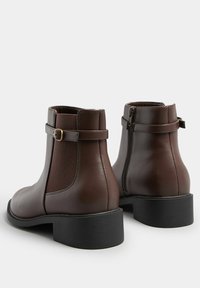 Botas de cuero marrón hasta el tobillo con cremallera lateral, panel elástico y una correa decorativa. Con un tacón negro bajo y texturizado.