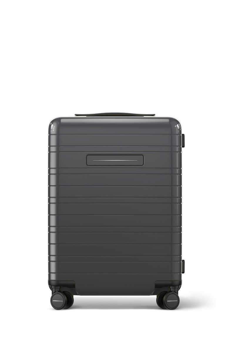 Valise rigide grise avec des rainures horizontales, une poignée supérieure et quatre roues pivotantes pour une maniabilité aisée. Dispose d'une fente pour une étiquette d'identification sur le côté.