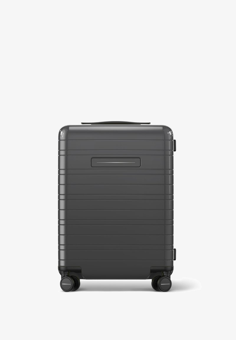 Valise rigide grise avec des rainures horizontales, une poignée supérieure et quatre roues pivotantes pour une maniabilité aisée. Dispose d'une fente pour une étiquette d'identification sur le côté.