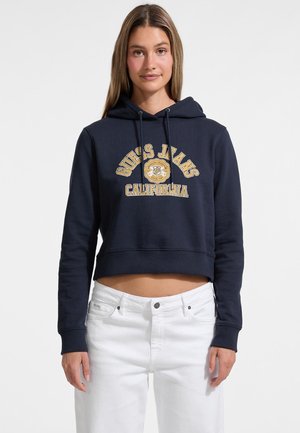 Navy cropped hoodie met trekkoorden, voorzien van het 'Guess Jeans California' logo in gouden letters. Gecombineerd met witte jeans.