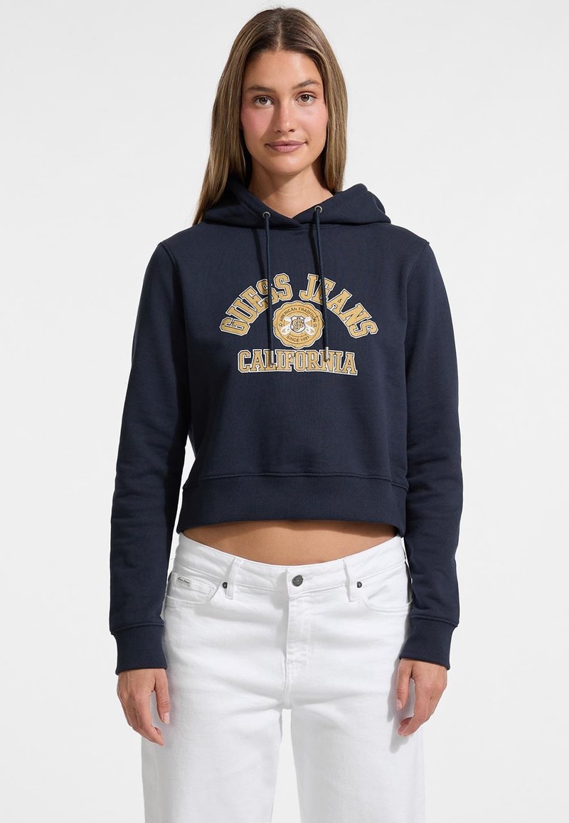 Felpa cropped blu navy con coulisse, caratterizzata dal logo "Guess Jeans California" in lettere dorate. Abbinata a jeans bianchi.