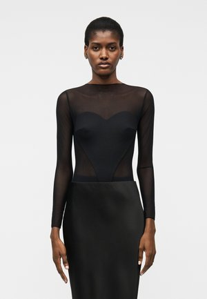 Sötét bőrű, rövid hajú nő fekete, áttetsző hosszú ujjú felsőt visel szív alakú mellrésszel és fekete szoknyát, egyszínű háttér előtt áll.