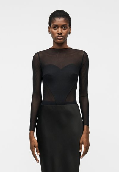 Femme à la peau foncée et aux cheveux courts portant un haut noir transparent à manches longues avec un corsage en forme de cœur et une jupe noire, debout devant un fond uni.