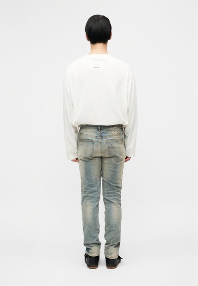 Valge pika varrukaga särk ja tuhmunud sinised teksad. Teksadel on kulunud tekstuur koos peente metalldetailidega tagataskutel. Mustad kingad.