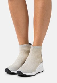 Bottes stretch métalliques dorées à la cheville avec une finition texturée, présentant une semelle blanche et une semelle extérieure en caoutchouc noir. Accents bleu clair en haut.