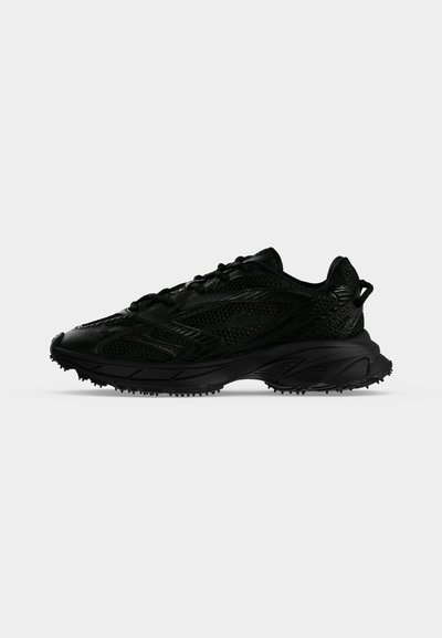 Chaussure de sport noire avec maille texturée et superpositions synthétiques, semelle épaisse avec crampons et languette au talon, vue de côté sur fond blanc.