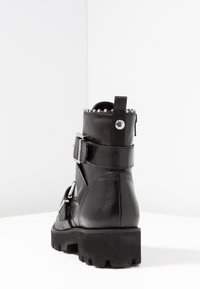 Stivale ankle in pelle nera con suola spessa, cinturini con fibbia argentata, trim borchiato in alto e linguetta con logo metallico su una superficie bianca.