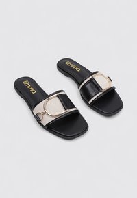 Sandalias planas negras con una amplia correa que presenta un acento circular dorado y un panel beige texturizado. Materiales duraderos y un diseño de puntera cuadrada.