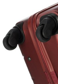 Angolo della valigia rigida rossa che mostra il materiale testurizzato, la cerniera liscia e due ruote girevoli nere per la manovrabilità.