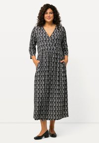 Mustapukinen mekko, jossa on valkoinen abstrakti kuvio, kolmivartiset hihat, sivutaskut ja liehuva helma. Yhdistetty mustiin korkokenkiin.