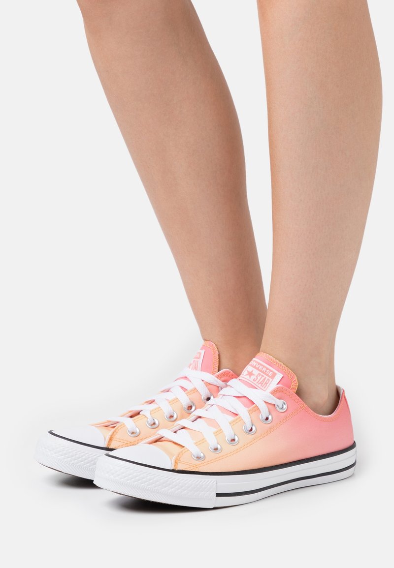 Różowo-pomarańczowe sneakersy kanvasowe z białymi sznurowadłami, gumowym czubkiem i czarną paskowaną podeszwą. Zaokrąglony kształt, klasyczny design.