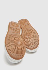 Para białych trampek z brązowymi gumowymi podeszwami z okrągłymi i liniowymi wzorami bieżnika, pokazanych od spodu.