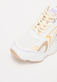 Scarpa da ginnastica bianca con sezioni in rete, dettagli gialli e lilla, linguetta imbottita e suola testurizzata. Design con lacci e occhielli rinforzati.