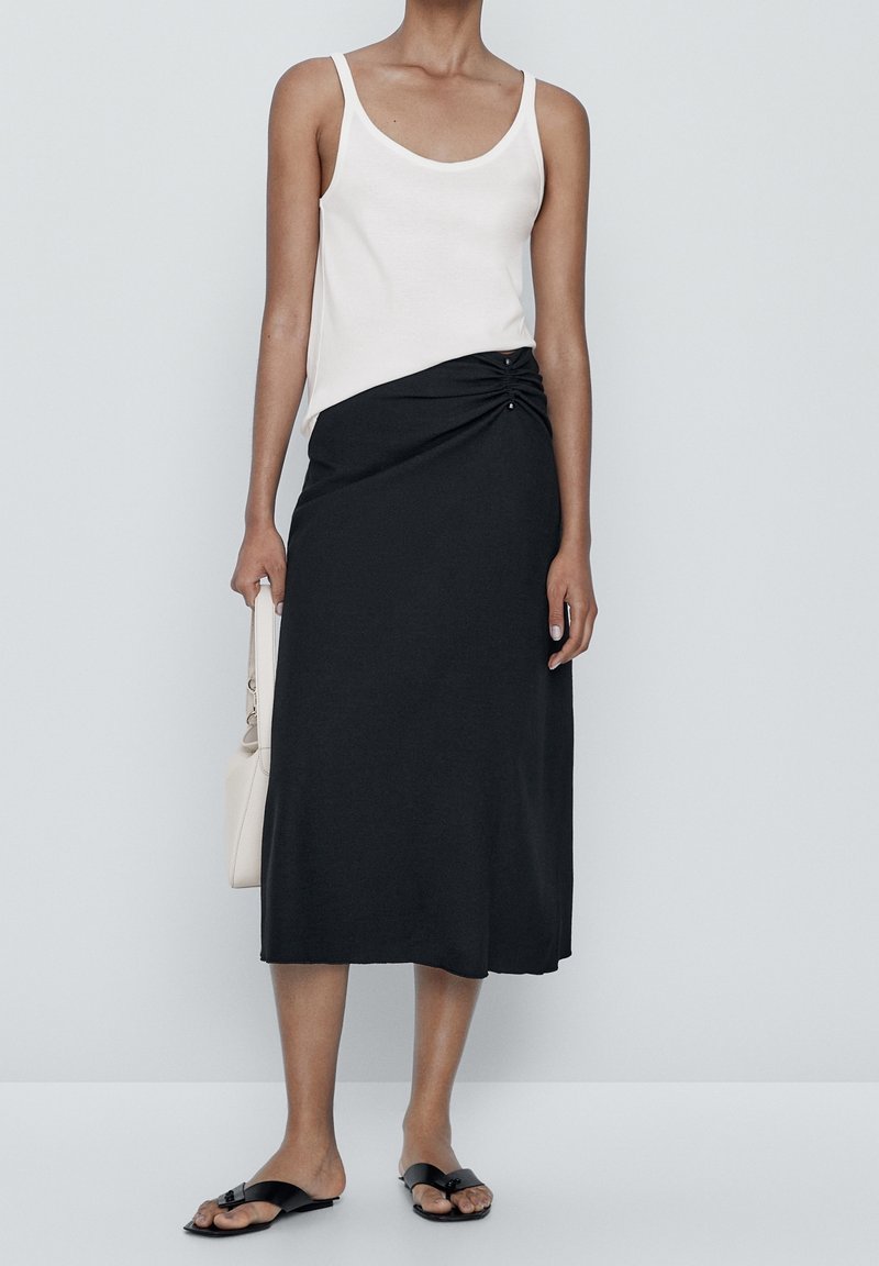 Femme portant un haut blanc sans manches et une jupe midi noire avec fronces sur le côté, tenant un sac à main beige et portant des sandales plates noires.