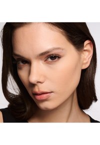 Primo piano di un viso con capelli lisci e scuri, pelle liscia, trucco naturale, occhi marroni e un leggero tocco di colore sulle labbra. Sfondo minimalista.