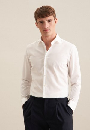 Jeune homme portant une chemise blanche cintrée rentrée dans un pantalon noir taille haute, debout les mains dans les poches devant un fond neutre.