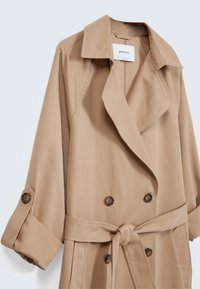 Beige double-breasted trench coat with wide lapels, tortoiseshell buttons, rolled-up sleeves, and a tied waist belt.