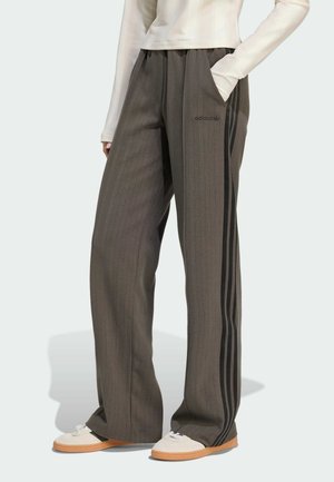 Personne portant un pantalon large marron Adidas avec des bandes latérales noires, main dans la poche, associé à des baskets blanches et un haut à manches longues clair.