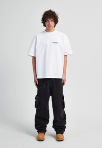 Weißes Oversized-T-Shirt mit dem aufgedruckten Logo "RETENITY", kombiniert mit schwarzen Cargo-Shorts mit Taschen. Beige Arbeitsschuhe runden das Outfit ab.