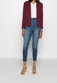 Femme portant un blazer bordeaux, un t-shirt blanc, un jean taille haute bleu avec des poignets retroussés, et des sandales à talons ouverts blanches, debout sur un sol gris.