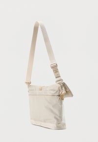 Μπεζ τσάντα χιαστή με ρυθμιζόμενο λουρί, εξωτερική τσέπη με φερμουάρ και κλείσιμο με τύλιγμα στην κορυφή, σε απλό ανοιχτό φόντο.