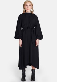 Rochie neagră, cu mâneci lungi, gât înalt, talie răsfrântă și siluetă fluidă. Prezentă cu mâneci bufante și o curea legată. Pur tată cu bocanci negri până la gleznă.