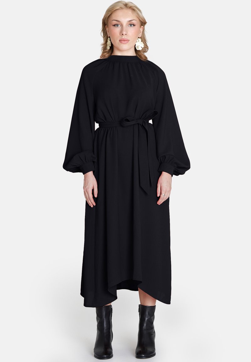 Rochie neagră, cu mâneci lungi, gât înalt, talie răsfrântă și siluetă fluidă. Prezentă cu mâneci bufante și o curea legată. Pur tată cu bocanci negri până la gleznă.