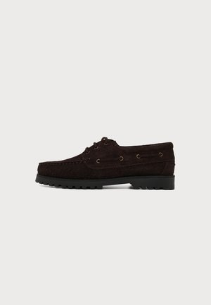 Chaussure bateau en daim marron foncé avec lacets à l'avant, couture moc toe, œillets latéraux et semelle en caoutchouc noire à crampons sur fond blanc.