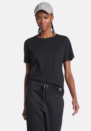 Femme portant une casquette grise, un t-shirt noir à manches courtes et un pantalon noir avec cordon, debout devant un fond clair uni.