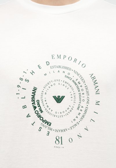 Λευκό ύφασμα με συγκεντρικούς κυκλικούς χαρακτήρες που αναγράφουν «Emporio Armani Milano Established 1981» και ένα κεντρικό στυλιζαρισμένο λογότυπο αετού σε πράσινο χρώμα.