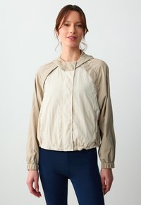 Beige Leichtjacke mit Kapuze, ausgestattet mit elastischen Bündchen und einem cinchten Saum. Glatte Textur und kontrastierende Farbpanels.