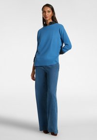 Blauer Strickpullover mit hohem Kragen, kombiniert mit weit geschnittenen Jeans aus Denim. Die Jeans sind in Blau, aus strukturiertem Stoff, mit einem klaren, klassischen Design.