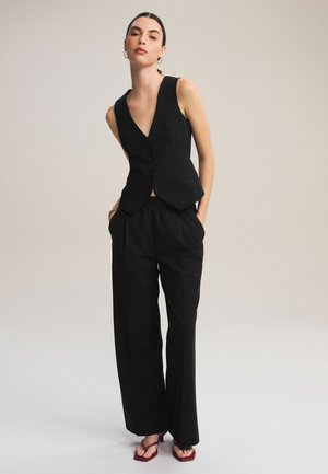 Nő fekete, ujjatlan, gombos mellényt visel, széles szárú fekete nadrágot és bordó szandált, zsebeibe dugott kézzel áll bézs háttér előtt.