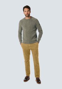Grau gestrickter Pullover mit geripptem Saum, kombiniert mit sandfarbenen Cordhosen und braunen Schnürschuhen. Das Model steht vor einem hellen Hintergrund.