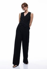 Zwarte vest met zes knopen, gecombineerd met hoge taille, brede zwarte broek. Kenmerken zijn strakke lijnen en een getailleerde pasvorm. Elegant en gestructureerde stof.