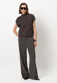 Grijze gebreide top met korte mouwen en een verhoogd golfpatroon, gecombineerd met grijze wijde broek. Model is accessorized met zwarte zonnebril.