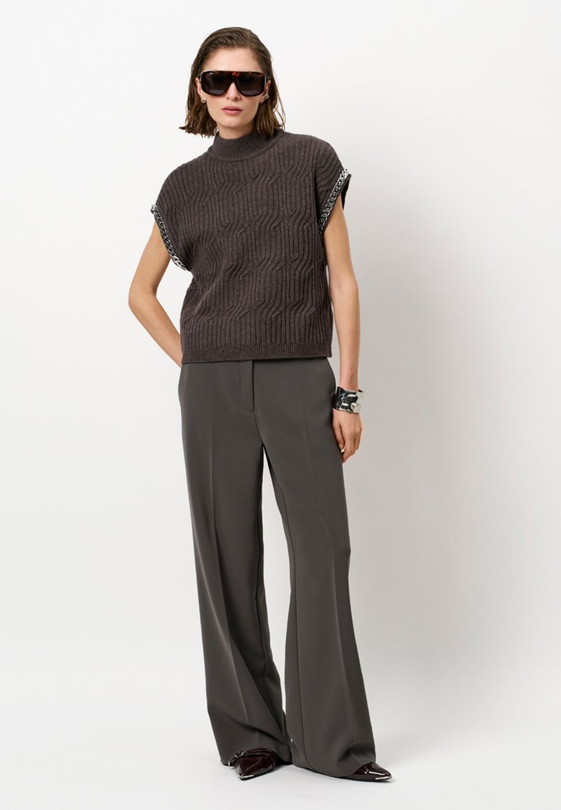 Grijze gebreide top met korte mouwen en een verhoogd golfpatroon, gecombineerd met grijze wijde broek. Model is accessorized met zwarte zonnebril.