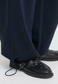 Mocassins en cuir tressé noir avec boucle argentée, portés avec un pantalon large bleu marine doté d'une sangle ajustable au niveau de la cheville.