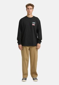 Junger Mann steht frontal und trägt ein schwarzes Langarmshirt mit einem blauen und rosa Wellenlogo, beige Hosen und schwarze Sneakers.