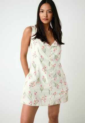 Frau mit langen dunklen Haaren, die ein ärmelloses weißes Kleid mit rosa und grünen Blumenmuster und Knöpfen vorne trägt, Hände in den Taschen.