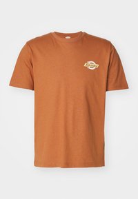 Camiseta de algodón color óxido de manga corta y cuello redondo, con un pequeño logo en el lado izquierdo del pecho y un dobladillo recto.