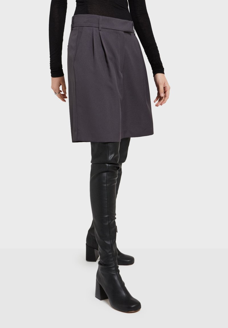 Grijze hoge taille geplooide rok, ontworpen in een gestructureerde stof. Gecombineerd met zwarte leren laarzen tot boven de dijen en robuuste hakken.