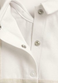 Polo blanc avec col à boutons. Présente des boutons-pression en argent et une texture en coton doux. Détails de couture visibles près du bord.