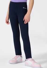 Leggings azul marino de corte ajustado, fabricados con tela elástica, con un pequeño logo blanco de "Champion" en el muslo. Llevados con zapatillas rosas y blancas.